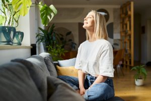 Happy woman sitting on a sofa with eyes closed, enjoying natural daylight indoors